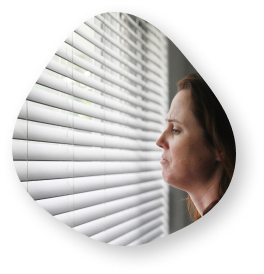 A woman looking out through window blinds with a worried expression, symbolizing anxiety and concern.