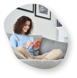 A young woman sitting comfortably on a gray couch, smiling and using a smartphone in a cozy living room.