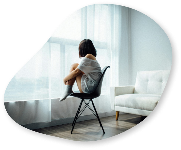 A woman sitting on a chair by a large window, looking outside with her knees drawn to her chest, appearing to be deep in thought.