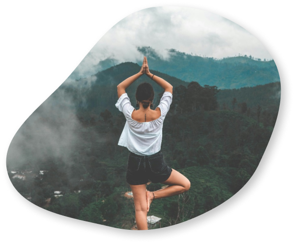 A woman practicing yoga on a mountain top, standing in a tree pose with her hands in a prayer position above her head, surrounded by misty hills.