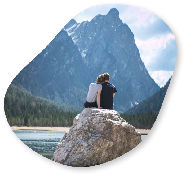 A couple sitting on a large rock by a lake, with towering mountains in the background, sharing a quiet moment together.