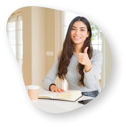 A woman sitting at a white table with a notebook, smiling and giving a thumbs-up gesture, symbolizing positivity and approval.