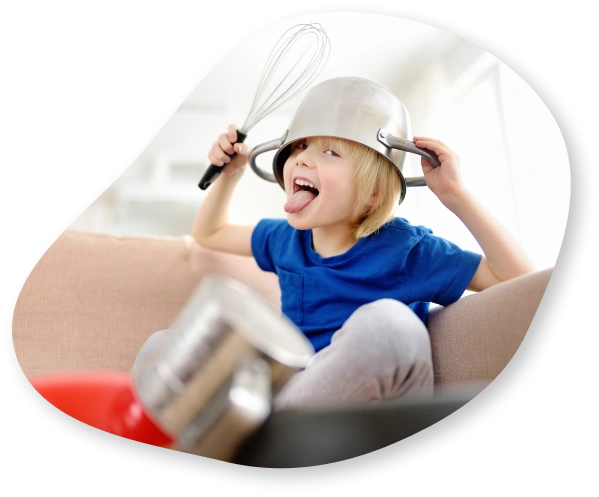 A young child with a whisk in his hand, a pot on his head, and a playful expression with his tongue sticking out.