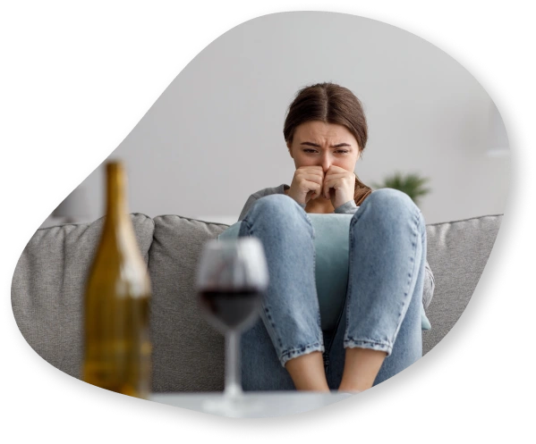 A woman sitting on a couch with both hands in fists, covering her mouth. She appears stressed, and a glass of wine is on the table in front of her.