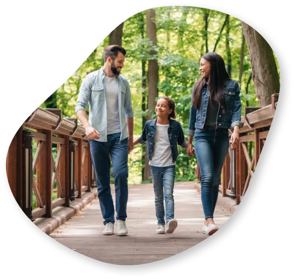 A family of three, a father, mother, and daughter, walking on a wooden bridge in a lush green park, smiling and enjoying their time together.