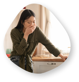 A woman standing in a brightly lit kitchen hunched forward. One of her hands is being used to lean on the counter while the other covers her mouth.